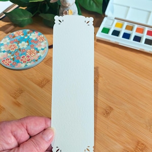 May include: A white bookmark with a decorative, scalloped edge. The bookmark is made of a textured paper and is blank.