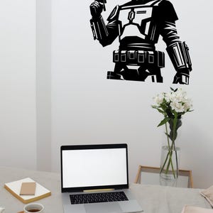 Peut inclure: Un autocollant mural noir représentant une illustration graphique stylisée d'un guerrier casqué en armure tenant un pistolet blaster. Le design montre un personnage avec une visière en forme de T, un plastron, une ceinture utilitaire et des gantelets.