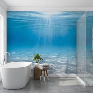 Op de afbeelding: Een moderne badkamer met een groot wit bad, een houten krukje met handdoeken en een plant in een pot. De muur toont een levendige blauwe onderwater scène met zonlicht. Een glazen douchecabine bevindt zich aan de rechterkant.