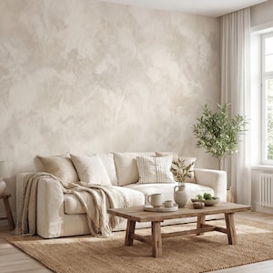 May include: A neutral-toned living room with a cream-colored sofa, throw pillows, and a woven blanket. A wooden coffee table displays decorative items. A jute rug and a small olive tree add warmth to the space. The wall has a textured, light-colored finish.