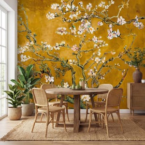 Puede incluir: Un comedor con una mesa redonda de madera y cuatro sillas de mimbre. La pared de color mostaza presenta un mural de un árbol en flor con flores blancas y rosas. Las plantas en macetas y una alfombra tejida completan la decoración.
