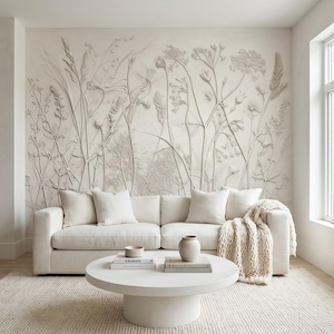 Op de afbeelding: Een witte bank met vier kussens en een grofgebreide plaid staat voor een muur met een groot reliëf-muurornament van wilde bloemen. Op een ronde witte salontafel staan twee stapels boeken en een kleine keramische vaas.