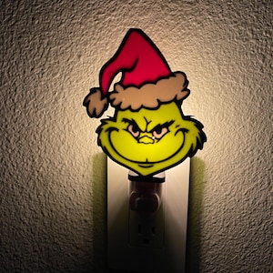May include: A novelty night light depicting the Grinch's face wearing a Santa hat. The Grinch's face is green with a yellow glow, and the hat is red with a brown trim. The night light plugs into a standard wall socket.