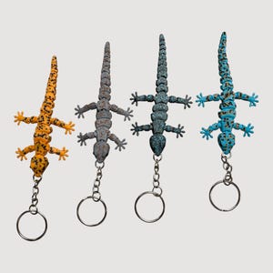 May include: Four lizard-shaped keychains in orange, gray, dark teal, and blue. Each lizard has a textured body and is detailed with black spots. The keychains are attached to silver key rings.