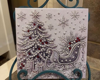 May include: A square canvas art piece featuring a Christmas tree and a sleigh decorated with white pearl-like beads and small red gems. The design includes snowflake patterns and gift boxes, all displayed on a teal metal scrollwork stand.