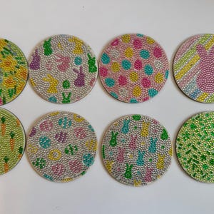 May include: Eight round, colourful coasters with various Easter-themed designs. The coasters feature a variety of patterns, including bunnies, carrots, eggs, and flowers. The coasters are made of a shiny, sparkly material.