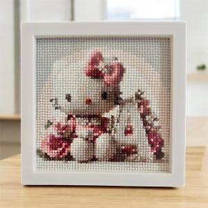 Puede incluir: Una pintura de diamantes enmarcada en blanco, de forma cuadrada, con un personaje de dibujos animados con un lazo y una bufanda rojos. La obra de arte está compuesta por pequeñas gemas coloridas y brillantes, creando un efecto de mosaico.