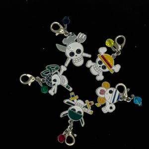 May include: Five metal charms featuring cartoon skull designs with various accessories. Each charm has a lobster clasp and a decorative bead. The charms are white with colorful accents, including green, yellow, and blue.