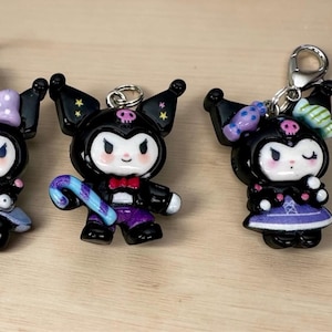 May include: Five black and white character keychains with silver clasps. Each figure has a skull on its head and is wearing different outfits, including a dress, bow, and hat. The keychains are on a light brown surface.