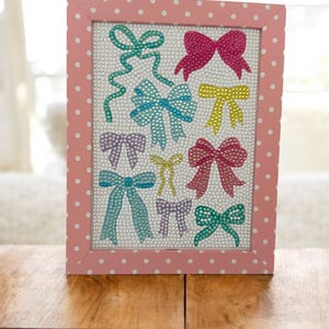 May include: A pink polka-dotted picture frame displaying a collection of colorful bow designs. The bows are embellished with sparkling details, set against a white background. The frame is resting on a wooden surface.