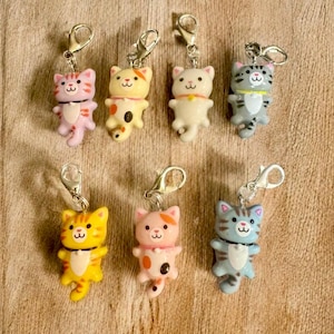 Puede incluir: Una colección de siete llaveros con forma de gato en varios colores, incluyendo rosa, amarillo, blanco y gris. Cada llavero presenta un cierre plateado y una pequeña figura de gato de dibujos animados con una cara sonriente y detalles decorativos.