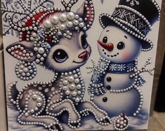 May include: A square canvas art piece featuring a cartoon deer with large eyes and a snowman wearing a top hat and scarf. The image is embellished with white pearl-like beads outlining the figures, snowflakes, and a small ornament on the ground.
