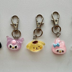 May include: Five character-themed keychains with silver-colored clasps. The charms are in the shape of cartoon characters, including a dog, a purple cat, a yellow dog, a pink bunny, and a clear character with a pink bow. The keychains are on a white background.