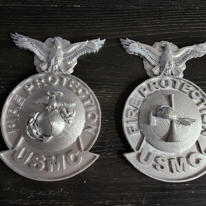 May include: Two silver-colored USMC Fire Protection badges. Each badge features an eagle at the top, a circular emblem with text, and the letters "USMC" at the bottom. One badge has a globe and anchor, the other a firefighter's helmet and axe.