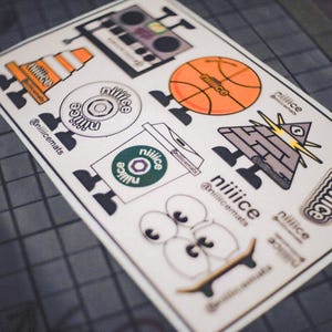May include: A sticker sheet featuring various cartoon graphics, including a basketball, boombox, traffic cone, and a pyramid with an eye. The stickers have the word "niiice" and the @niilicemats handle.