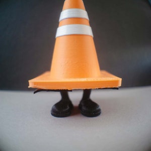 Op de afbeelding: Een kleine oranje verkeerskegel met witte strepen staat op twee zwarte benen. De kegel draagt zwarte schoenen.