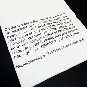 Könnte beinhalten: Weißes T-Shirt mit einem schwarzen Zitat von Michel de Montaigne, "Les Essais", Livre 1, Kapitel 26. Das Zitat lautet: "On demandait à Socrate d'où il était. Il ne répondit pas d'Athènes, mais : Du monde. Lui, qui avait son imagination plus pleine et plus étendue, embrassait l'univers comme sa ville, jetait ses connaissances, sa société et ses affections à tout le genre humain, non pas comme nous qui ne regardons que sous nous."