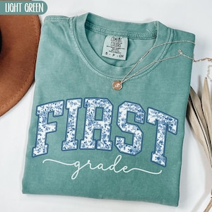 Może przedstawiać: Jasnozielona koszulka z napisem "FIRST grade" w niebieski kwiatowy wz&oacute;r. Koszulka jest złożona i ma metkę Comfort Colors. Złoty naszyjnik z wisiorkiem jest zarzucony na koszulkę.