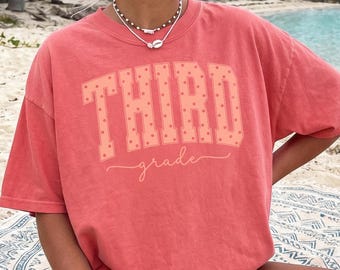 Könnte beinhalten: Ein korallenfarbenes Kurzarm-T-Shirt mit dem Text "THIRD grade" in hellem Pfirsichton. Das Wort "THIRD" ist in einer großen, gepunkteten College-Schriftart dargestellt, während das Wort "grade" in einer geschwungenen Schreibschrift geschrieben ist.