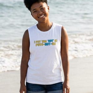 May include: White vest top with the text "I PUT THE HOT IN PSYCHO-NOT-IN" in yellow and blue. The person is wearing denim shorts and standing on a sandy beach with the sea in the background.