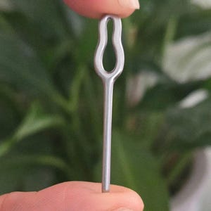 May include: A small, silver-colored metal pin with a looped top and a straight, slender stem. The pin is held between two fingers against a blurred green background, suggesting a craft or jewelry-making item.
