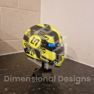 Peut inclure: Un casque de course jaune et noir avec un design noir et jaune comportant une lettre stylisée "L" et le mot "NORRIS". Le casque a une visière transparente et un autocollant bleu avec le texte "Tezos".