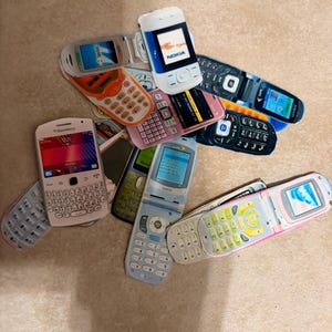 Peut inclure: Une collection de téléphones portables vintage, dont des téléphones à clapet et des smartphones. Les téléphones affichent une gamme de couleurs, notamment le blanc, le rose, le bleu et l'orange. Certains téléphones portent des logos de marques, comme Nokia et Blackberry. Les téléphones sont disposés en tas.