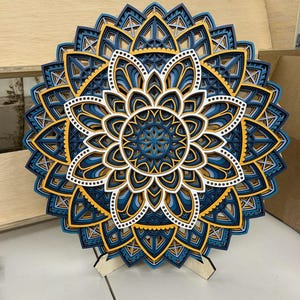 May include: A blue, white, and yellow layered mandala wall art with a wooden stand. The mandala has intricate geometric patterns and is made of wood.