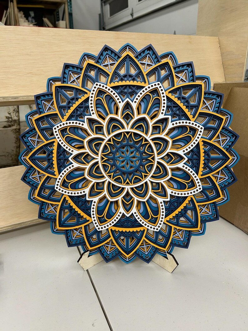 May include: A blue, white, and yellow layered mandala wall art with a wooden stand. The mandala has intricate geometric patterns and is made of wood.