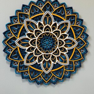 May include: A layered paper mandala with intricate designs in shades of blue, white, and yellow. The mandala is a circular design with a central flower shape and multiple layers of petals.