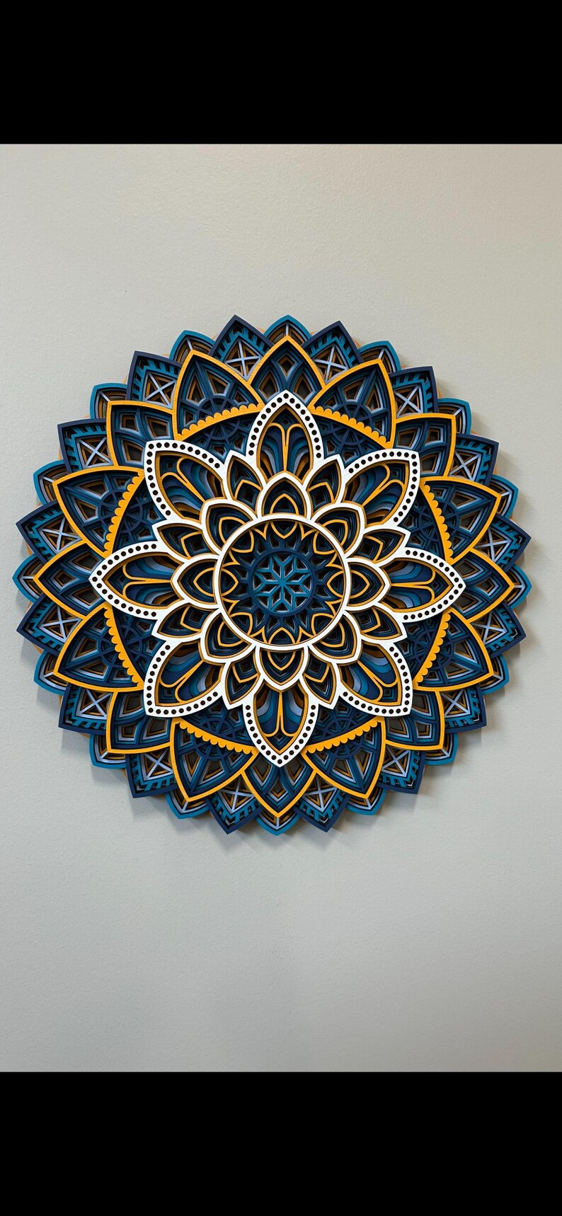 May include: A layered paper mandala with intricate designs in shades of blue, white, and yellow. The mandala is a circular design with a central flower shape and multiple layers of petals.
