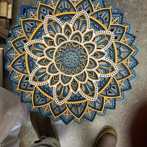 May include: A layered wooden mandala with intricate geometric patterns in shades of blue, yellow, and white. The mandala is circular and has a symmetrical design.