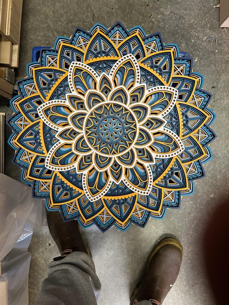 May include: A layered wooden mandala with intricate geometric patterns in shades of blue, yellow, and white. The mandala is circular and has a symmetrical design.