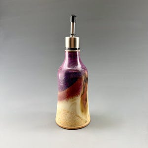 May include: A ceramic oil cruet featuring a tapered body with a glaze transition from deep purple and maroon at the top to a speckled beige and tan base. The vessel is topped with a stainless steel pourer spout and a small black plastic cap.