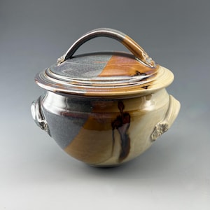 May include: A ceramic lidded pot with two side handles and a curved lid handle. The glaze features a split color design of dark charcoal gray and warm amber brown, with a smooth, glossy finish and subtle dark brown vertical drip patterns.