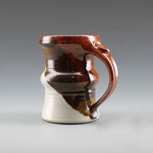 May include: Handmade ceramic mug with a unique design. The mug features a white base, transitioning to brown and reddish-brown hues. The handle is a matching reddish-brown. This stoneware mug is perfect for coffee, tea, or any beverage.