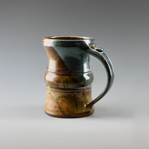 May include: A ceramic mug featuring a cylindrical body with a central horizontal ridge and a curved handle. The glaze displays a blend of earthy brown, tan, and deep blue tones, with a glossy finish that highlights the smooth, rounded contours.