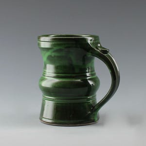 May include: A dark green ceramic mug with a glossy finish, featuring a cylindrical body with two horizontal raised bands around the center and a curved handle attached to the side. The glaze shows subtle variations in shade from deep to forest green.