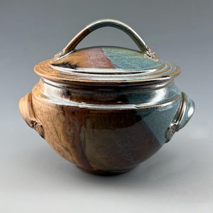 May include: A round ceramic lidded pot with two side handles and a curved handle on the lid. The glaze features a blend of earthy brown, deep black, and muted teal tones, with a glossy finish and visible variations in color across the surface.