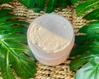 May include: A close-up of a white jar filled with a light pink and white exfoliating scrub. The jar is sitting on a woven straw surface.