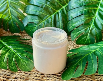 May include: A clear glass jar filled with a thick, white body butter. The jar is sitting on a woven brown surface with green leaves in the background.