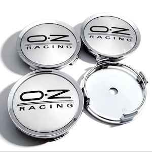 Puede incluir: Cuatro tapacubos plateados con el texto negro "O.Z RACING". Las tapas son redondas con una superficie brillante y reflectante. Una tapa está separada, revelando la parte trasera.