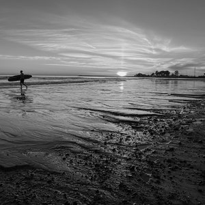 May include: A person walks along a sandy beach carrying a surfboard, with the sun setting in the distance.