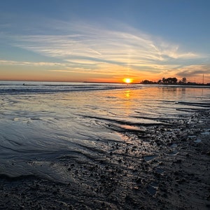 May include: A sunset over the ocean with a sandy beach in the foreground. The sky is a mix of orange, pink, and blue with wispy clouds. The water is calm and reflects the colors of the sky.