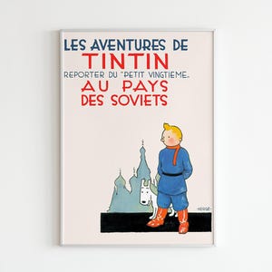 Op de afbeelding: Een ingelijste vintage poster met de titel "LES AVENTURES DE TINTIN" in blauw en rood. De poster toont een stripfiguur in blauwe kleding, rode laarzen en een witte hond, tegen een achtergrond van blauwe gebouwen. De tekst "AU PAYS DES SOVIETS" is ook zichtbaar.