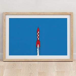 以下が含まれることがあります： 青い背景に赤いロケットが打ち上げられる様子を描いた額装されたイラスト