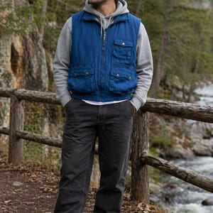 May include: A blue quilted gilet with multiple pockets, worn over a grey hooded sweatshirt and white t-shirt. The person is wearing dark grey trousers, brown boots, and a brown cap. The scene is outdoors near a wooden fence and a river.