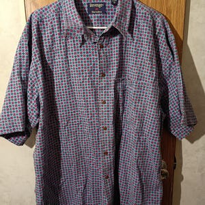 Puede incluir: Una camisa de manga corta con botones y un patrón repetitivo de cuadrados rojos y verdes sobre un fondo azul claro. La camisa tiene cuello, bolsillo en el pecho y la marca "Revenge" es visible.