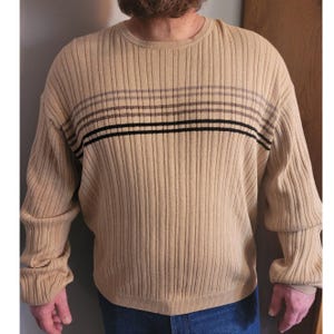May include: A tan ribbed knit sweater with horizontal stripes in black, brown, and grey. The long-sleeved sweater has a crew neck and a relaxed fit. The fabric has a vertical ribbed texture. This casual sweater is suitable for everyday wear.