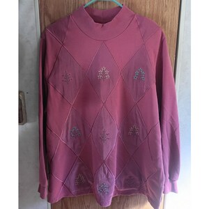 May include: A dusty rose long-sleeve pullover sweater featuring a mock neck collar and a quilted diamond pattern across the front. Each diamond section contains a small, delicate embroidered floral motif in shades of white and light blue.